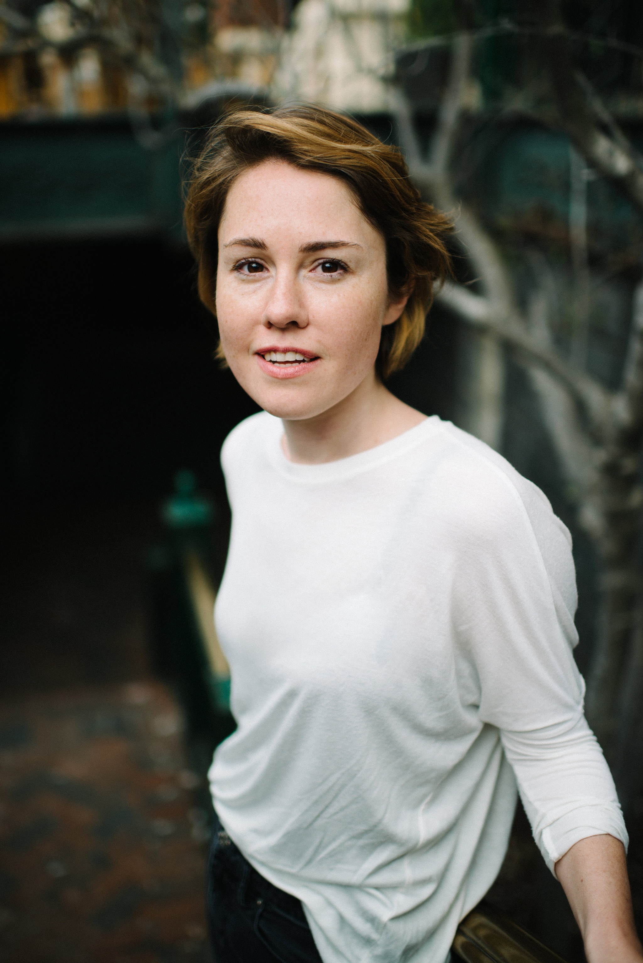 A white woman with short, light brown hair and wearing a sheer white blouse sits outside with bare branches in the background. 