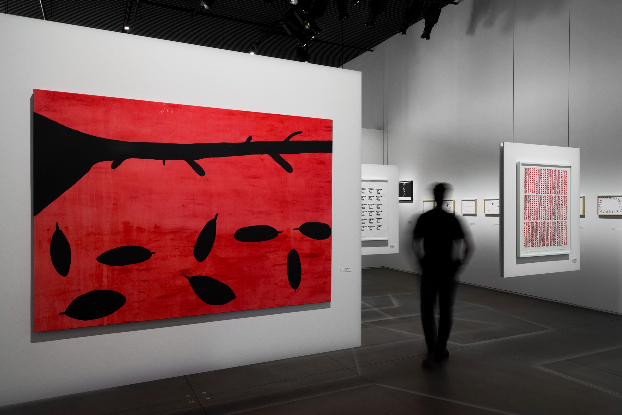 An art gallery with drawings and paintings hanging on fixed walls and suspended panels hanging from the ceiling. A visitor walks away between two suspended panels. In the foreground, is a rectangular painting hung on a horizontal orientation. It depicts bold black shapes that recall a fallen tree branch and large leaves against a bright red background. 