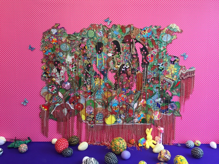 A colorful, multimedia tapestry is centered on a hot pink background with small white polka-dots, adorned with life-size butterfly sculptures. The tapestry features several Black teenagers and children casually interacting, conversing, and playing amongst oversized flowers and colorful toys and objects. Fringe and other adornments drape down over the work. The work extends from ceiling to floor. The floor is colored ultramarine and is scattered with brightly painted balls, many of which are egg-shaped, as well as stuffed animals.