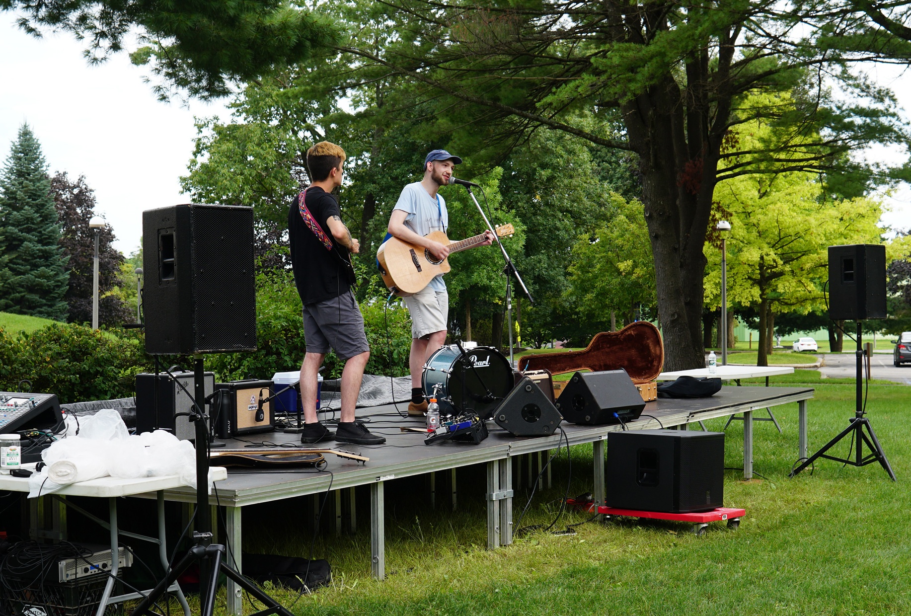 Two people stand atop an outdoor stage playing guitar, with lush green trees behind them. One sings into a microphone.