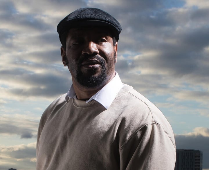 photo of a man wearing a black cap, beige sweater over a collared shirt with blue skies and clouds in background