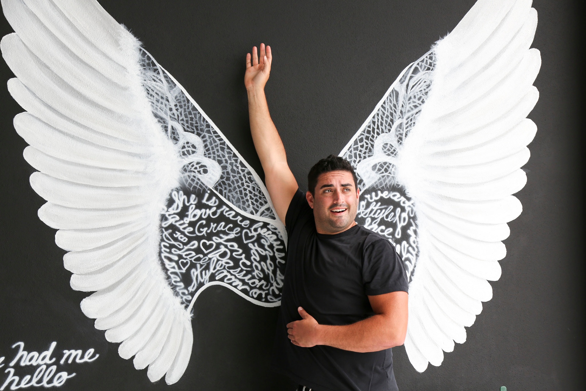 A man in a black t-shirt standing against a mural of white wings. He stands in the center of the wings where the meet, with one hand raised above his head. 