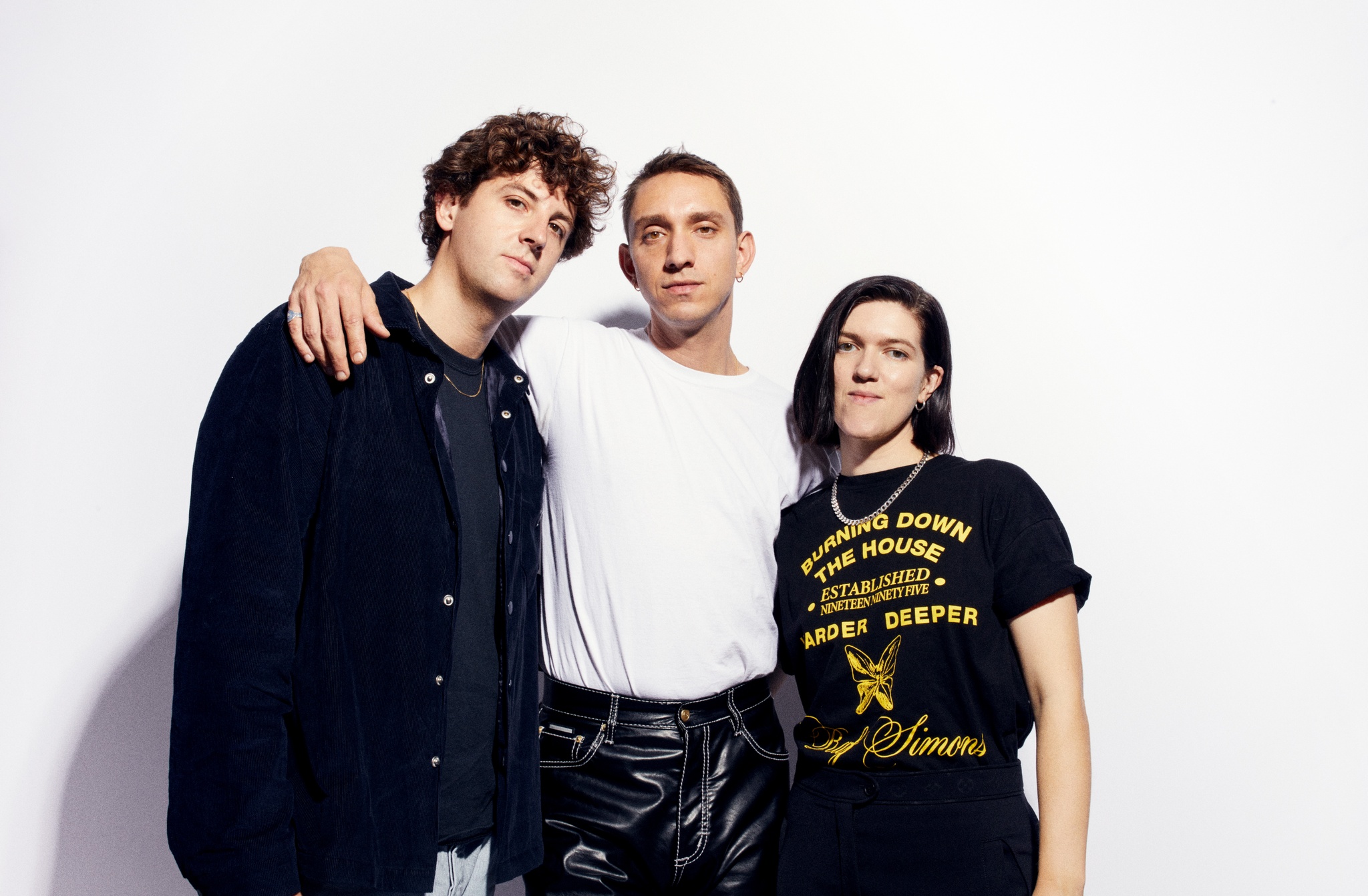 The three members of The xx standing against a white wall. One stands in the middle with his arms around the other two at either of his sides. The man in the middle wears a white shirt. On the left another man wears a dark shirt and jacket. On the right a woman with short brown hair wears a black tshirt. 