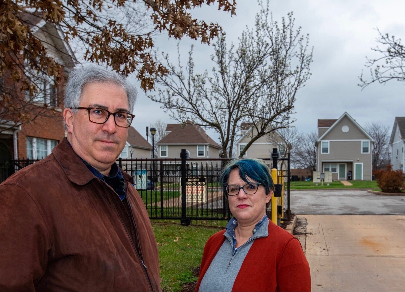 Two people standing in front of a residential area, with a black iron fence behind them, on a gray-sky day.