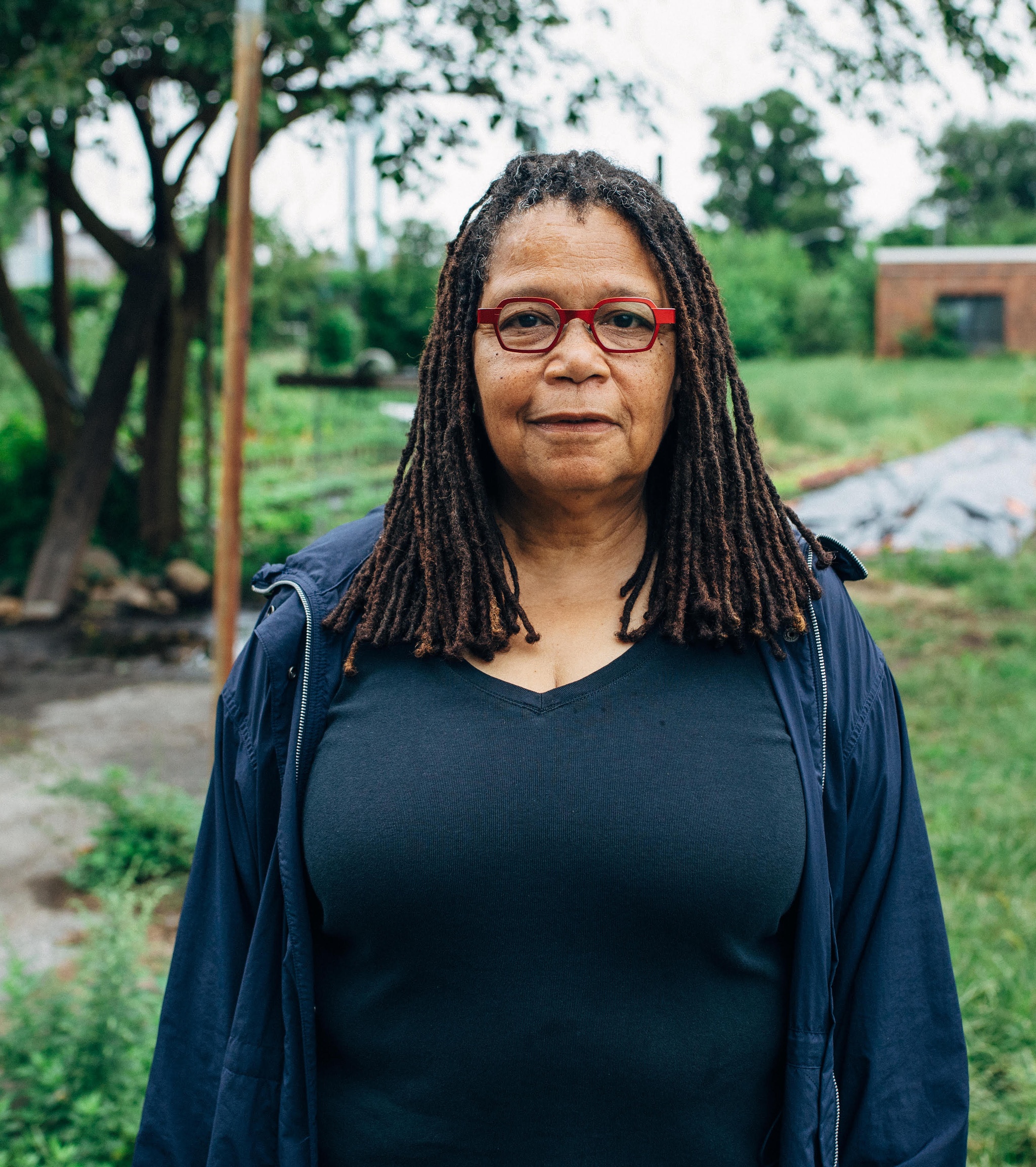 A Black woman with shoulder-length hair parted in the middle stands in front of a green, garden or backyard space. She wears red-framed glasses and smiles without opening her mouth.