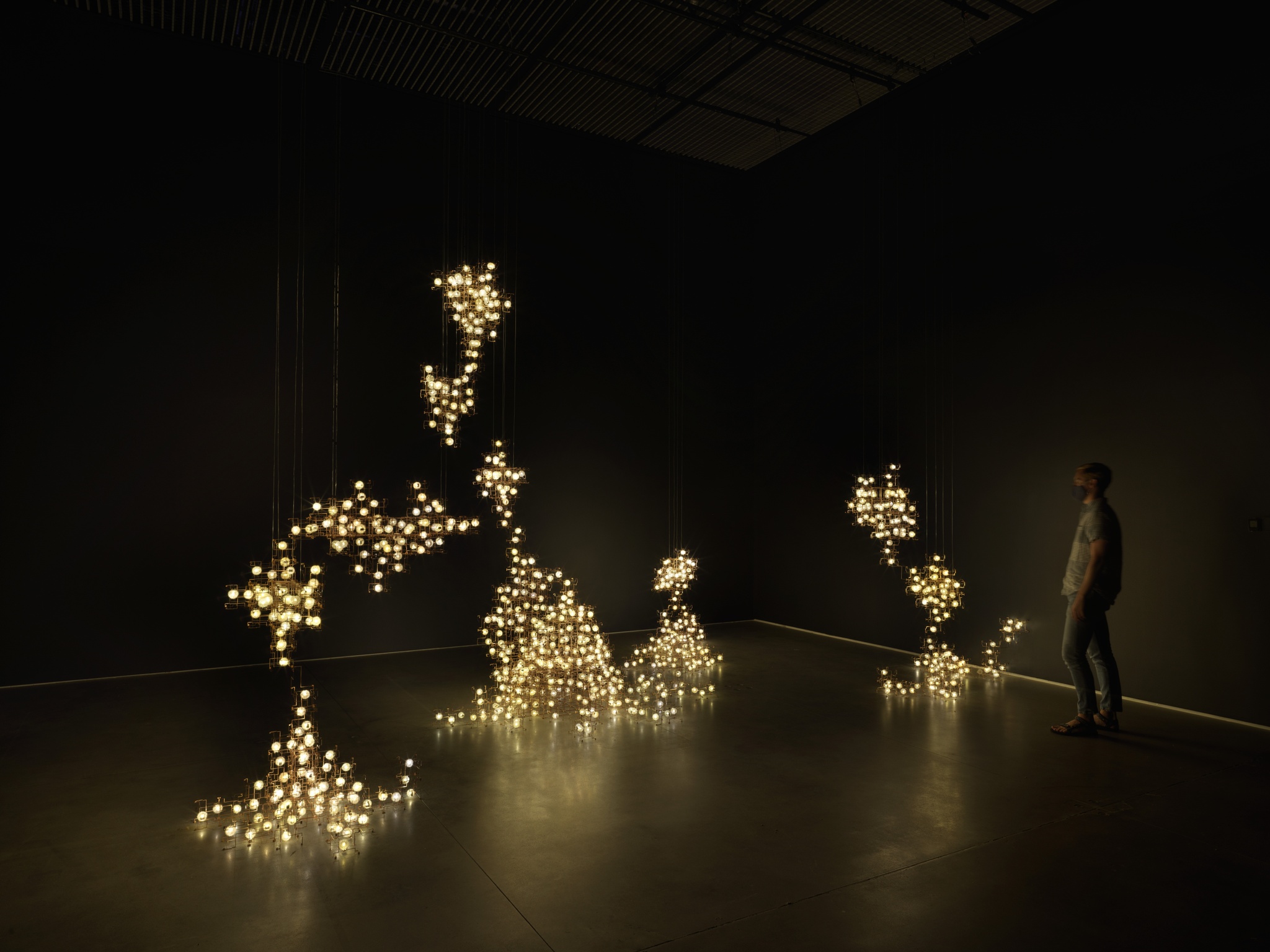 A person standing in a gallery space beside an installation of small twinkling lights arranged in a metal structure that seems to grow out of the floors and walls