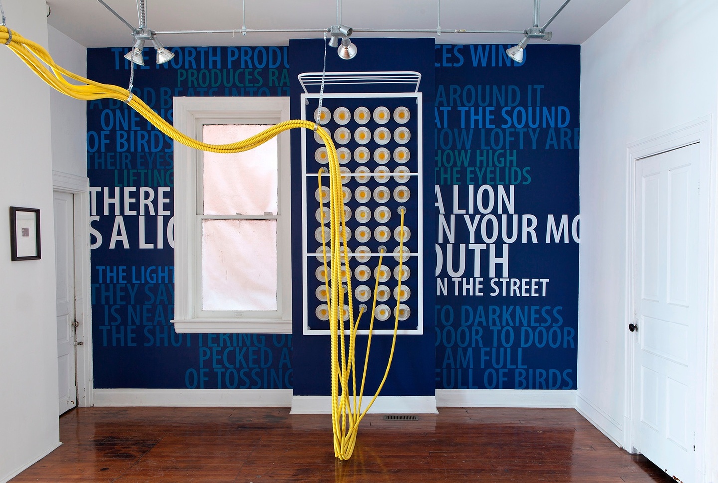 Installation view of a gallery space: yellow tubing hangs from the ceiling, suspended by white wires/cords, and connect to sockets where they plug into the wall. The walls are a deep ultramarine blue, with text of various colors (different shades of blue, teal, and white) tiled across. The walls to the left and right are white, and the floors are wooden.