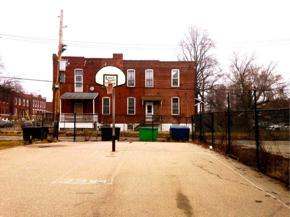 A basketball hoop on a court, with a red-brick building behind it. The words "Love Bank" are written on the backboard.
