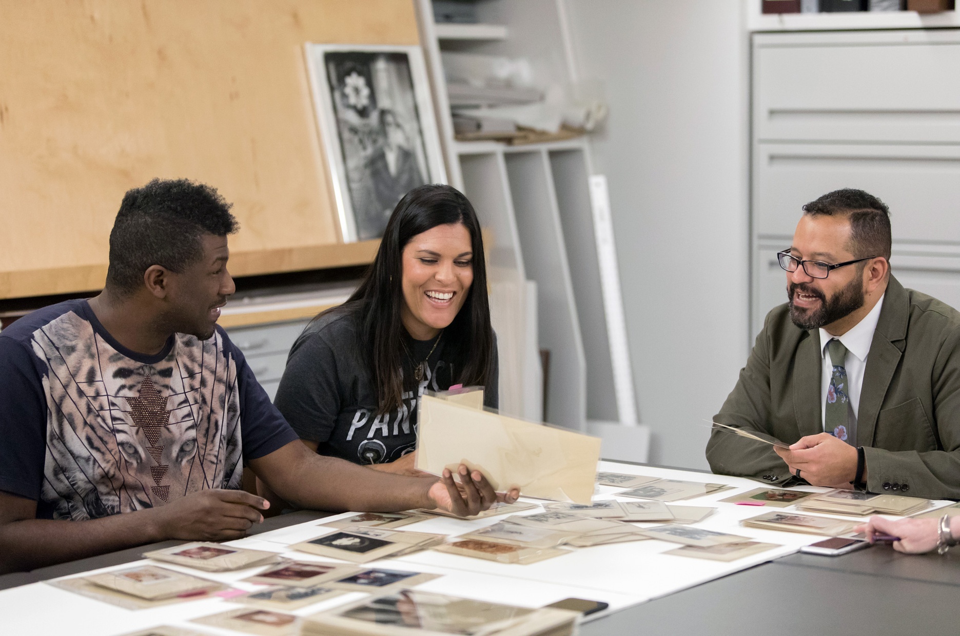 Three people sit around a table laughing and smiling looking at photographs laid out on the table.
