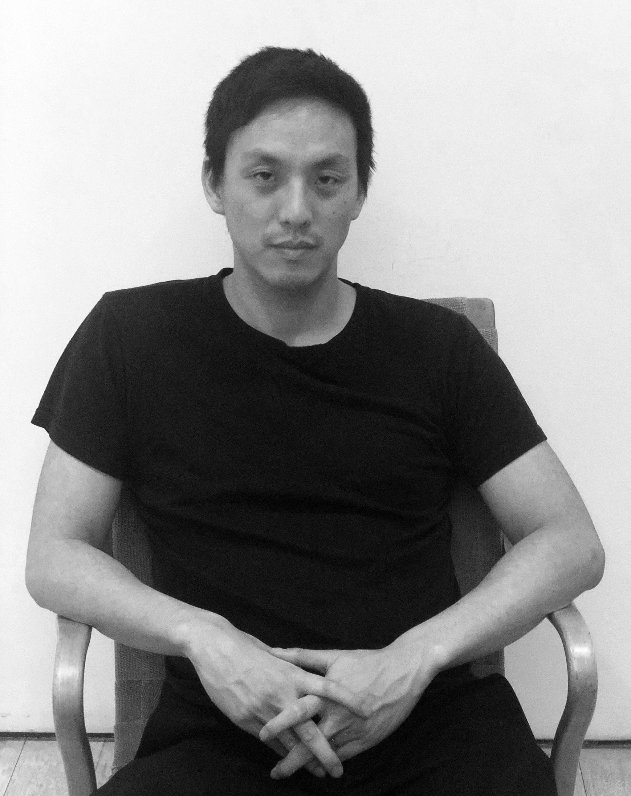 The artist Ian Cheng in a black t-shirt and pants, sitting in a chair with hands crossed in his lap