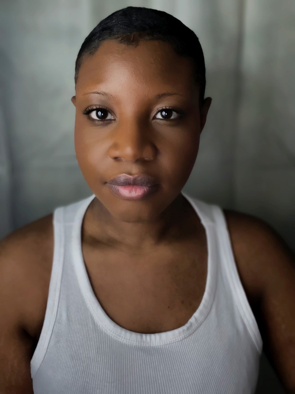 A headshot of actor Mara Allen, who looks directly at us. Mara has brown skin and wears a white tank top. 