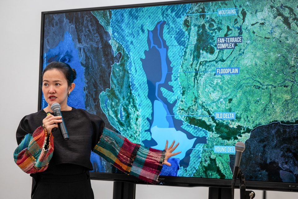 Voraakhom stands in front of a tv screen displaying an aerial map with an overlaid graphic showing floodplain borders.