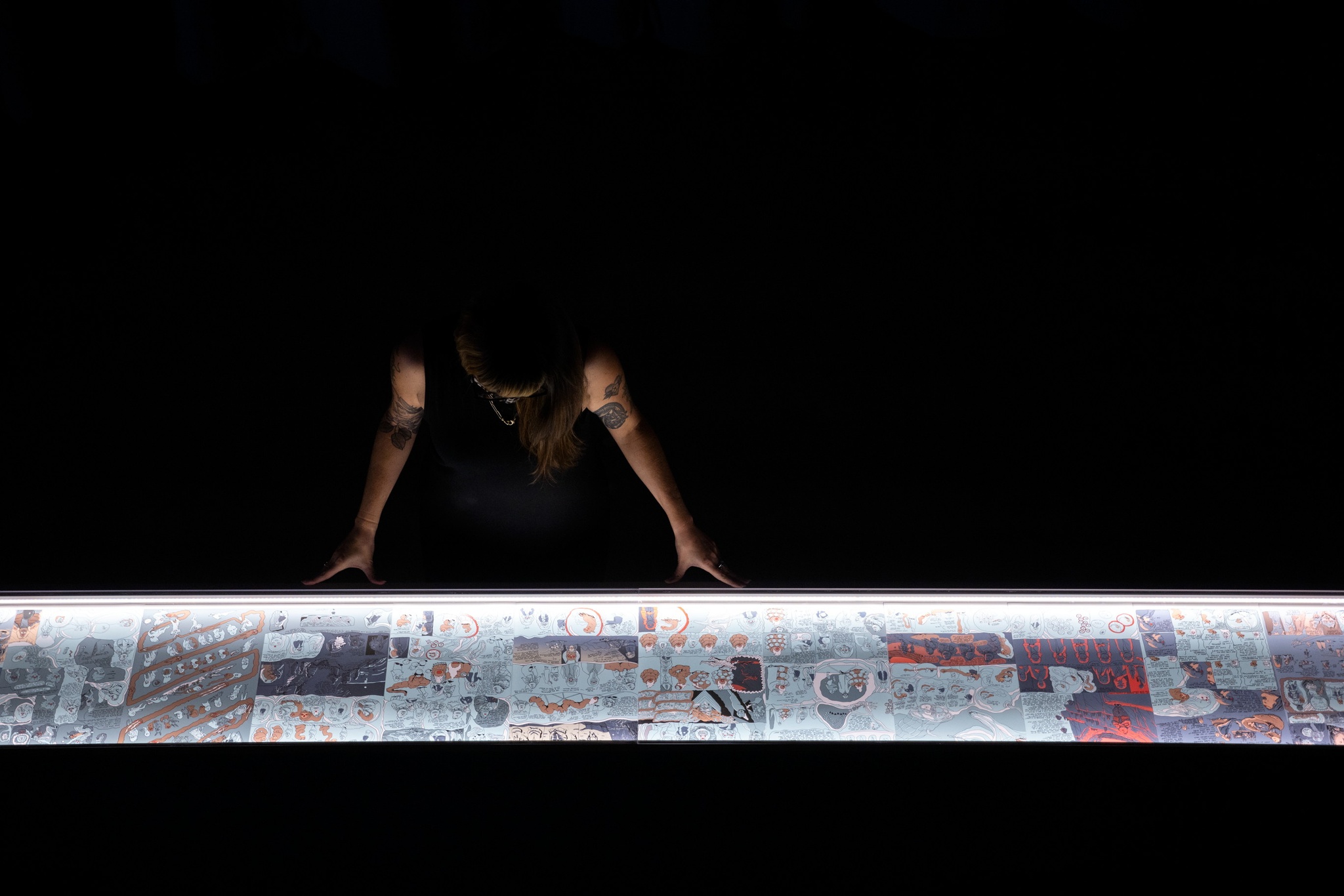 A person with tattooed arms leans out of the shadow over a light-box illuminating pages from a graphic novel
