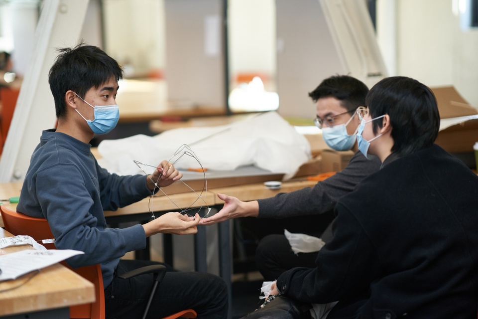 Three students sitting in a studio space look at a wire object.