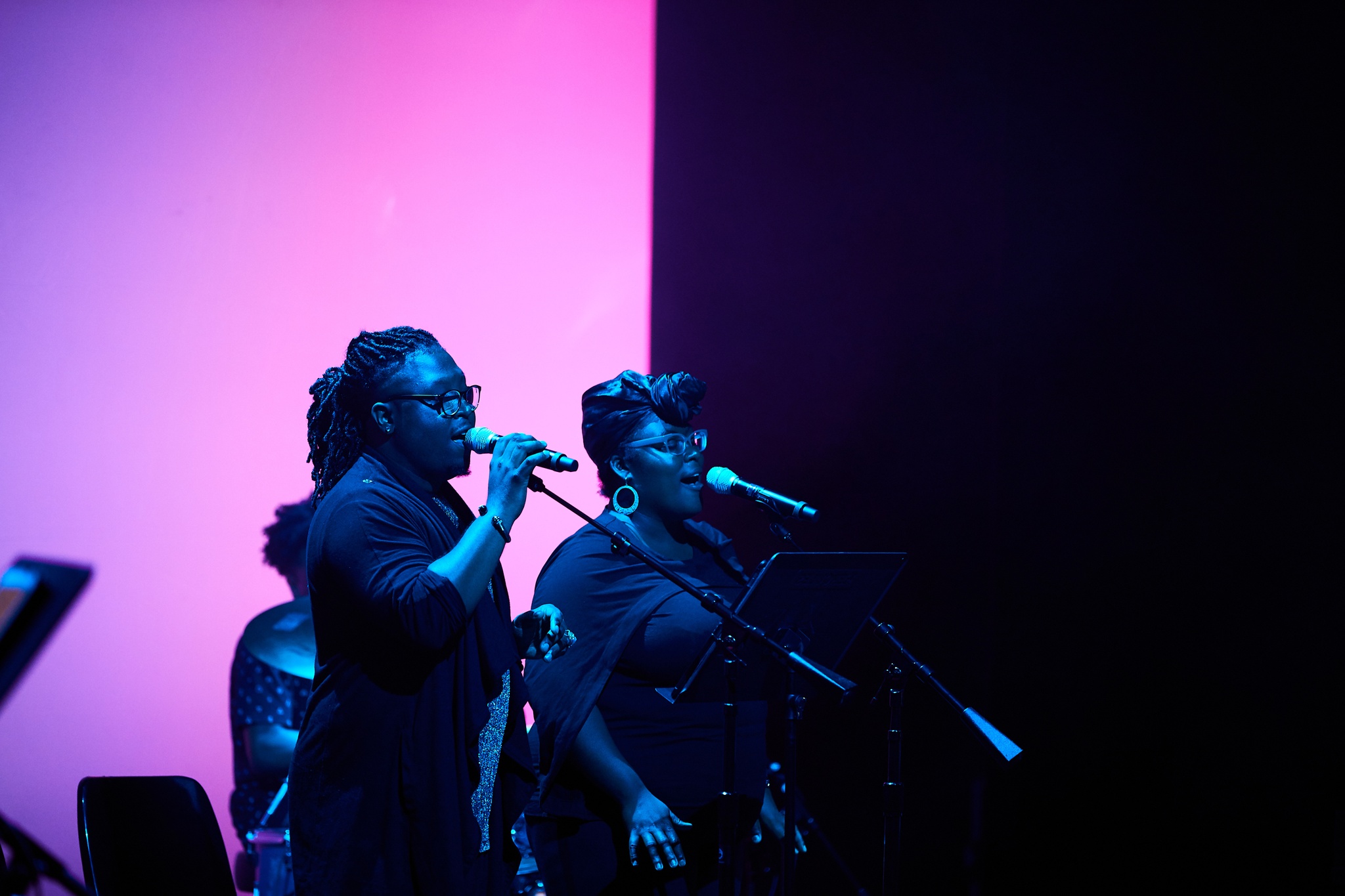 Two performers singing on blue-lit stage in front of large pink screen.