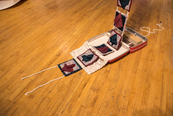 A vintage hardback suitcase sits open on the floor. A series of red, blue, and beige quilted fabric squares are attached in a vertical row, dangling over the suitcase and lying across it.