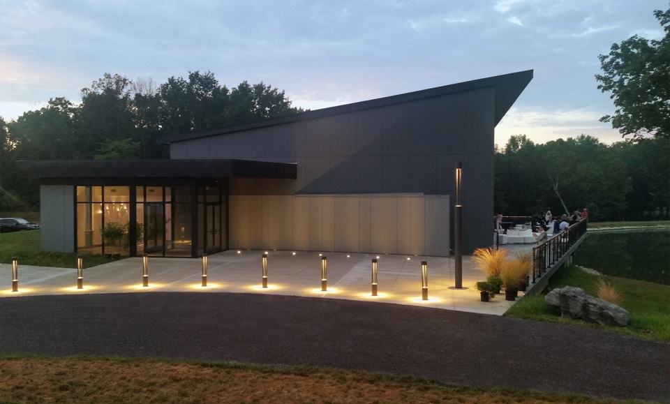 Photo of the exterior facade of a large banquet facility, situated on farmland, at dusk. The contemporary building has an angled, triangular roofline, with ground lighting in front.