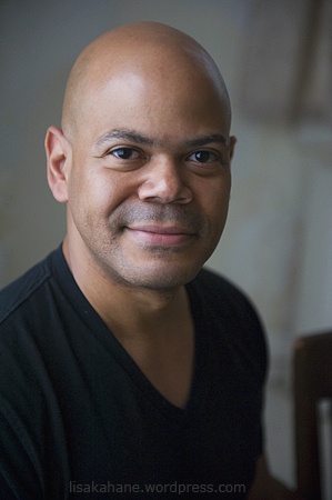 A headshot of Charles Rice-Gonzalez who has a clean shaven head and wears a v-neck tshirt. He smiles at us with a joyful look in his eyes. Photo by Lisa Kehane