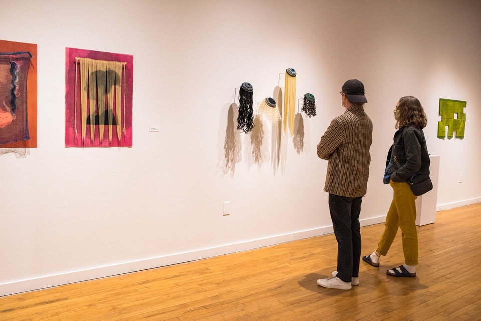 Two people in a gallery space look at a piece consisting of four wigs of different colors, textures, and lengths topped by yarmulkes projecting from the wall.