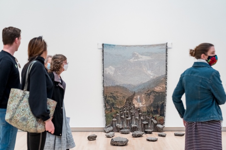 People walk past an installation of a woven blanket with the image of a hazy valley landscape on it. The bottom edge of the blanket has been picked apart and piece of asphalt have been hung from strings like beads. Larger chunks of asphalt litter the floor in front of it.