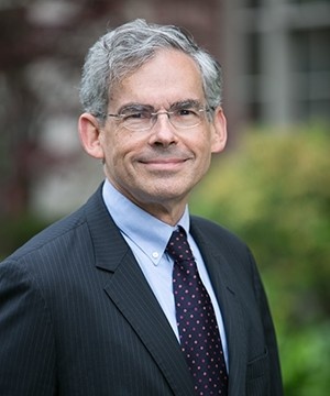 A white man with gray hair parted to the side smiles. He is wearing a suit and tie, as well as glasses, and standing outside with a green bush in the background.