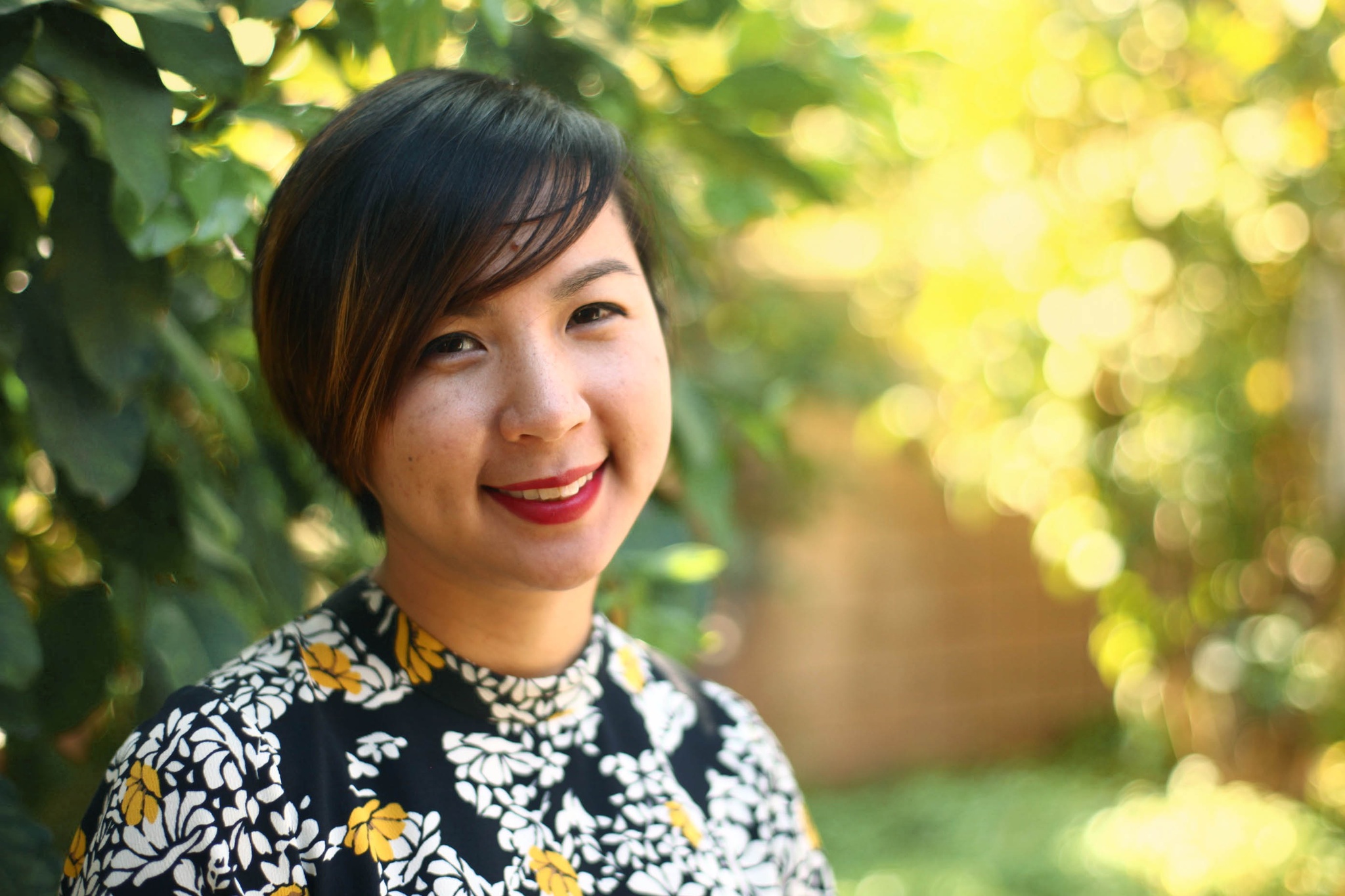 A headshot of Cathy Linh Che, a Vietnamese American woman who poses outside in sunlight with trees and bushes in the background. She has short brown hair swept across her forehead, wears red lipstick, and smiles at us. Photo by Jess X. Snow
