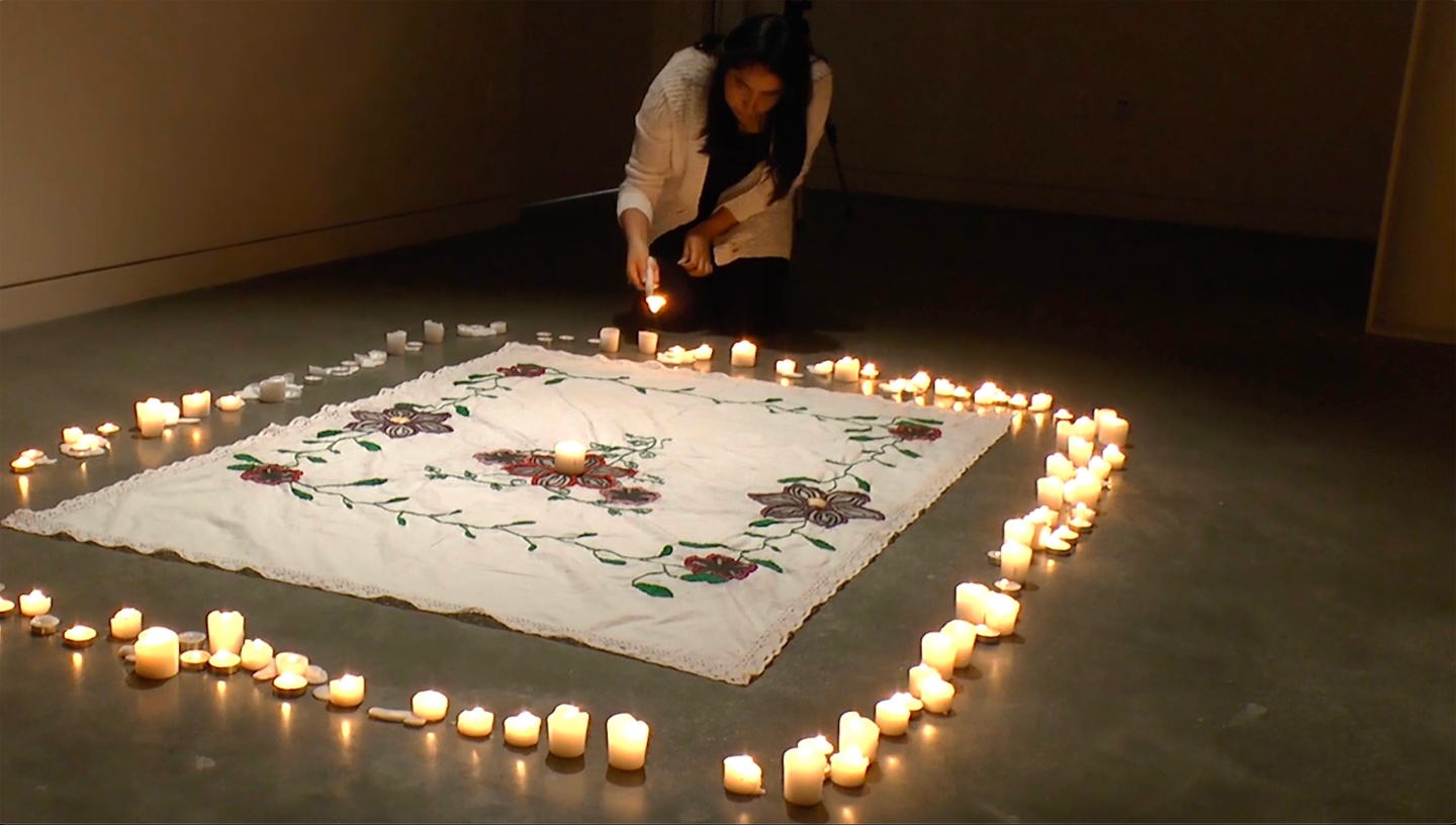 A person crouching down lighting candles, which surround a large rectangular embroidered piece of fabric in a spacious gallery. The fabric has lace detailing along the edges and is white. There is a candle placed in the center.