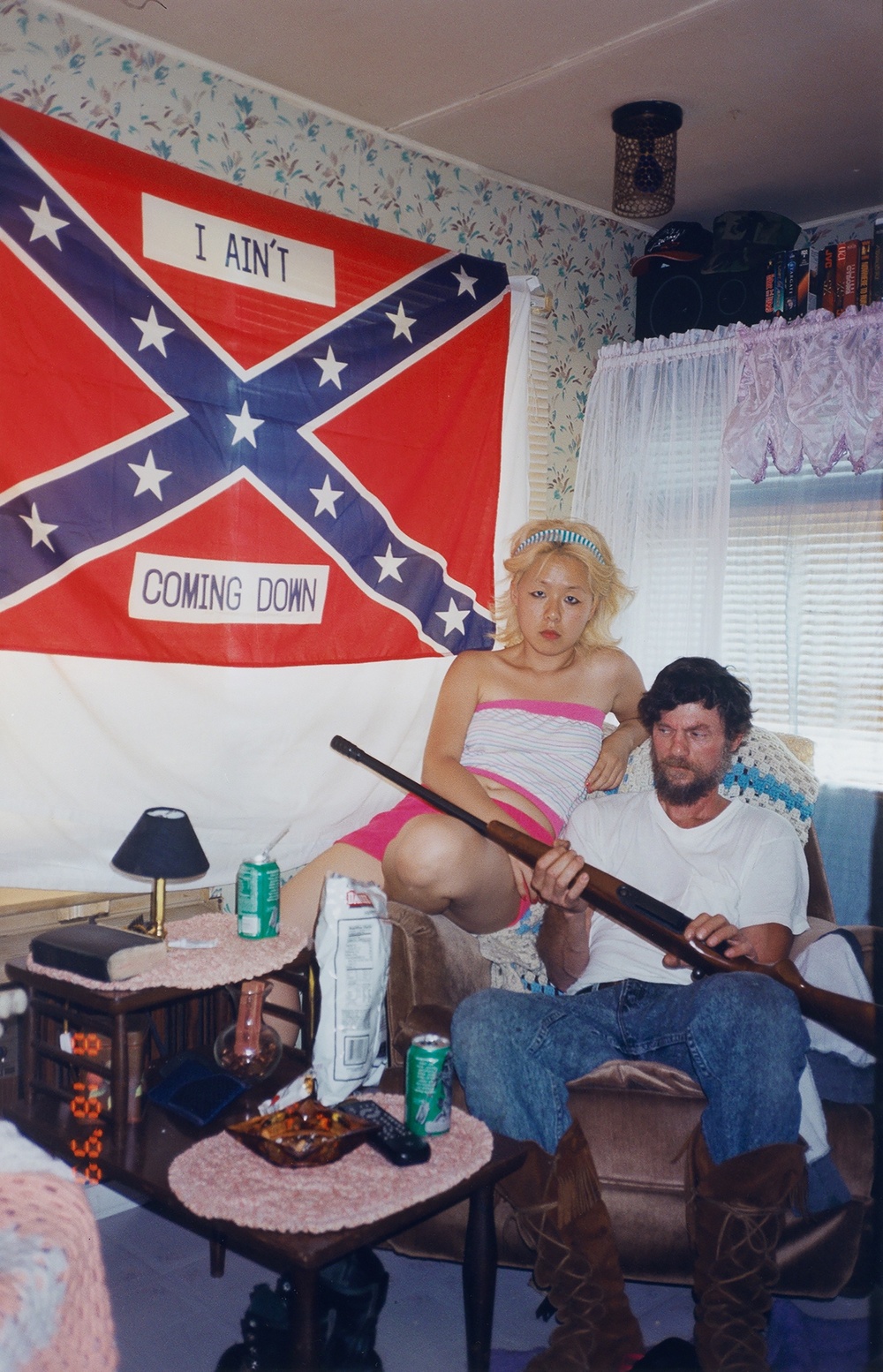In a living room hangs a confederate flag that reads “I AIN’T COMING DOWN,” below which sits a Korean woman with dyed-blonde hair next to a white male holding a rifle. 