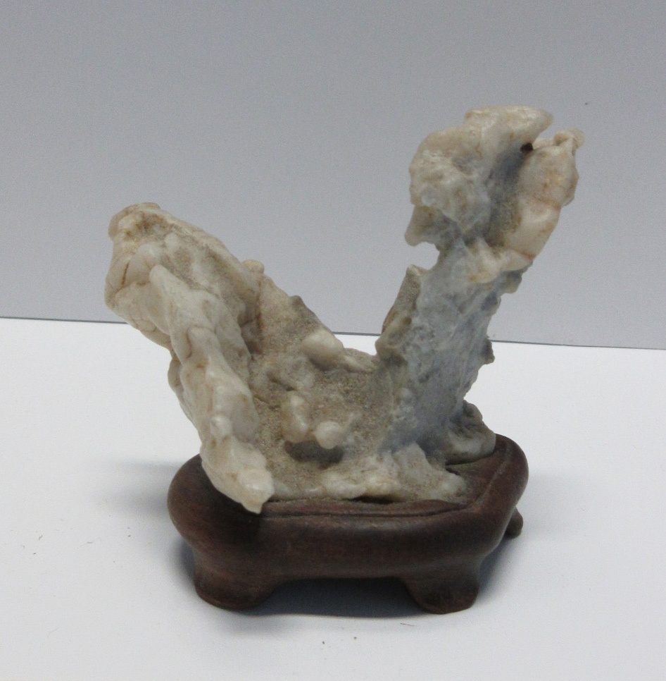 A lumpy naturally formed gray stone, with two diagonal offshoots pointing upwards in opposite directions forming a letter V shape, sits atop a dark brown wooden base.