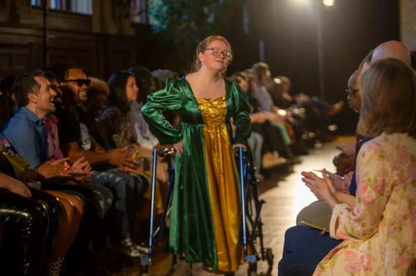 A young woman with a walker modeling a fairytale green and gold dress pauses on a fashion design show runway and smiles to an applauding audience.