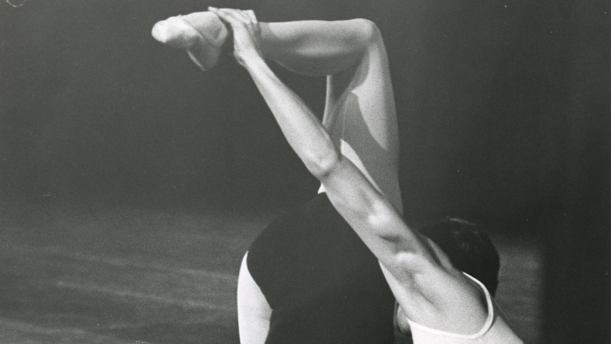 A black and white photograph of two ballet dancers, one balanced with body upside down and back to the camera, the other provides support at the head and raised foot.