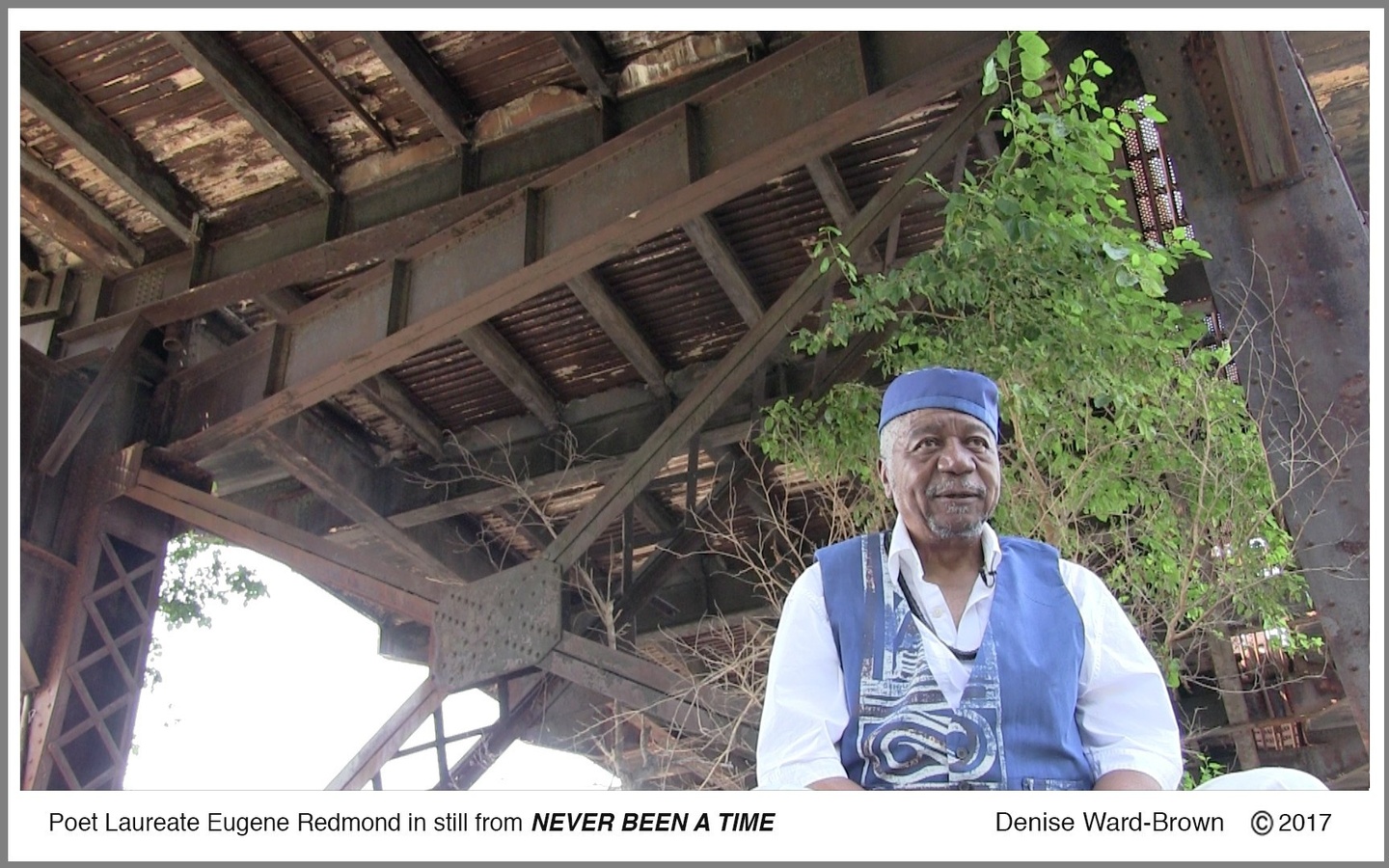 Text reads, "Poet Laureate Eugene Redmond in still from NEVER BEEN A TIME" above a still from the video, in which Redmond wears a white shirt with ea blue vest and cap, under the floor of a wooden structure. There is a tree behind him.