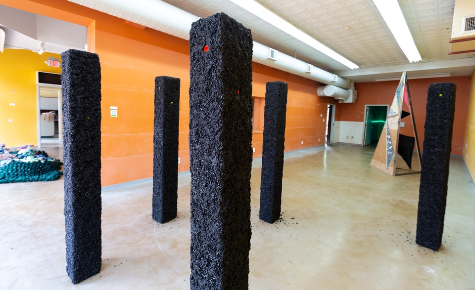 Gallery view of 5 asphalt covered pillars with small yellow and orange holes on them.