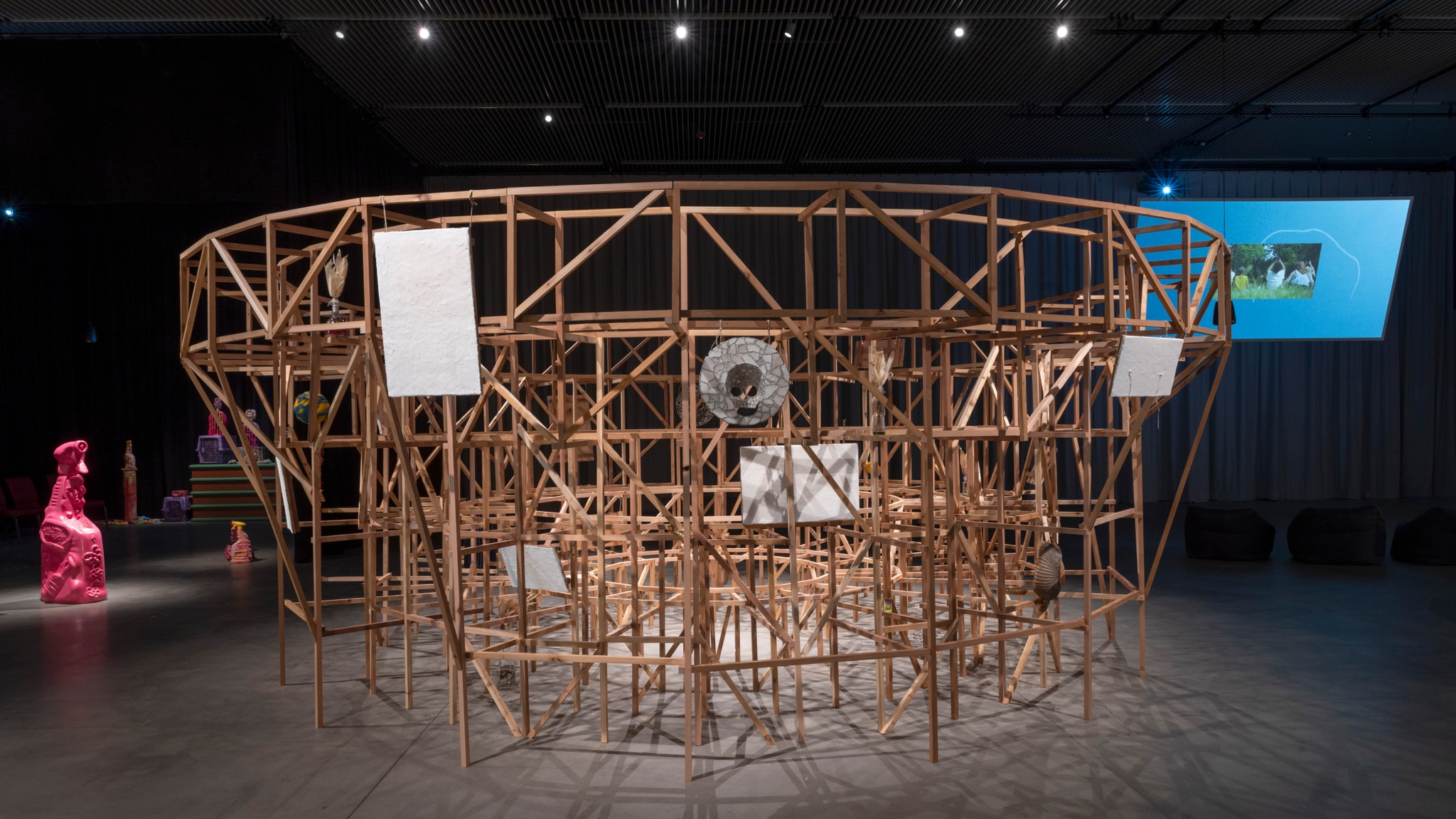 A wooden sculptural installation in a gallery with low lighting. The circular structure, about 6 feet high, is a scaled down model of the architectural support for arena seating. It is made of thin beams of wood and has objects like masks and paintings hanging from it facing outward. The center of the arena is empty and inaccessible to gallery visitors. 