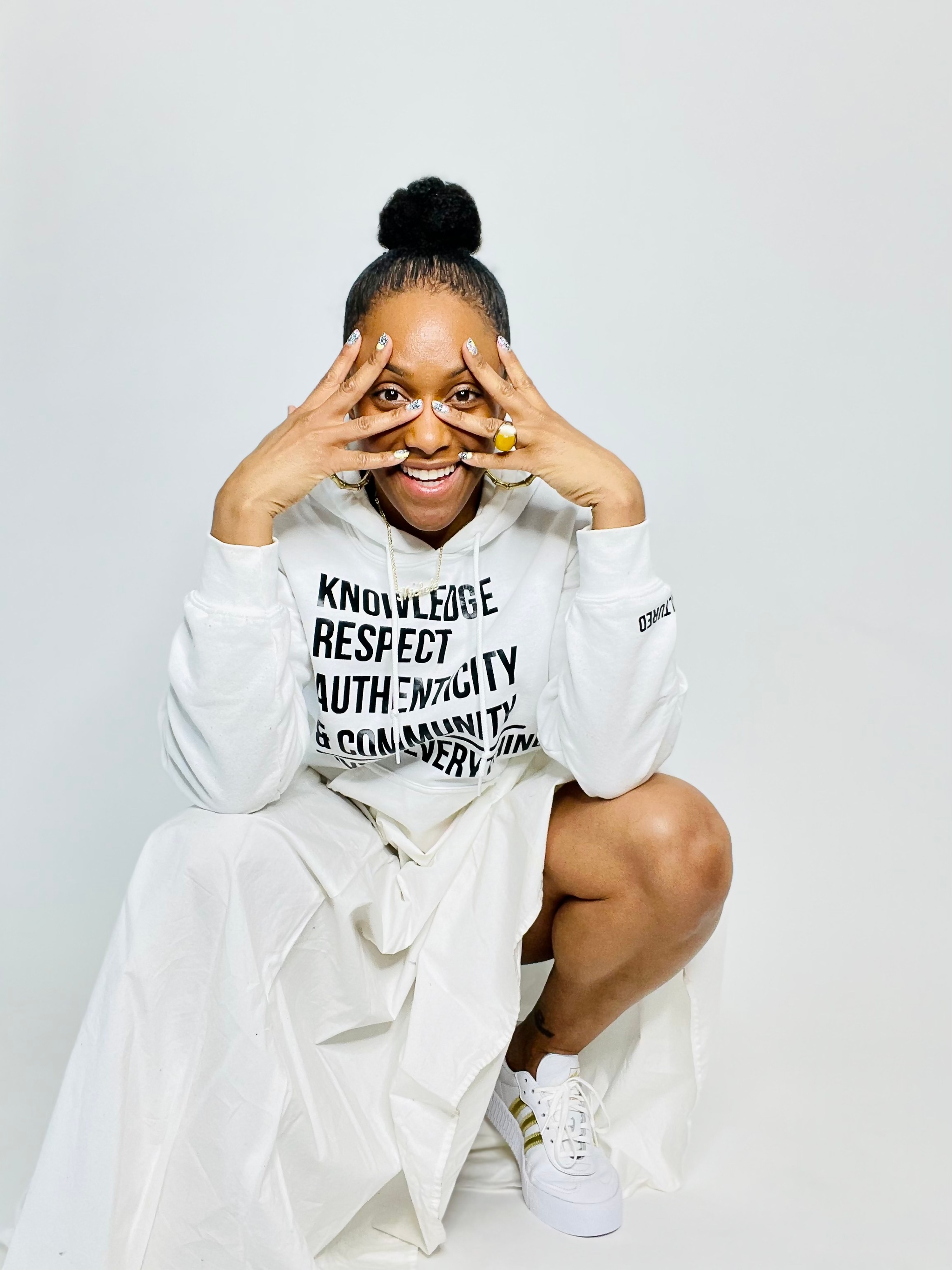 A portrait of dancer Michele Byrd-McPhee, a Black woman with light brown skin who crouches against a white background. She wears an all-white outfit with her hair in a round bun on top of her head. She smiles excitedly and holds her hands up to her face, peering through her fingers like a mask.