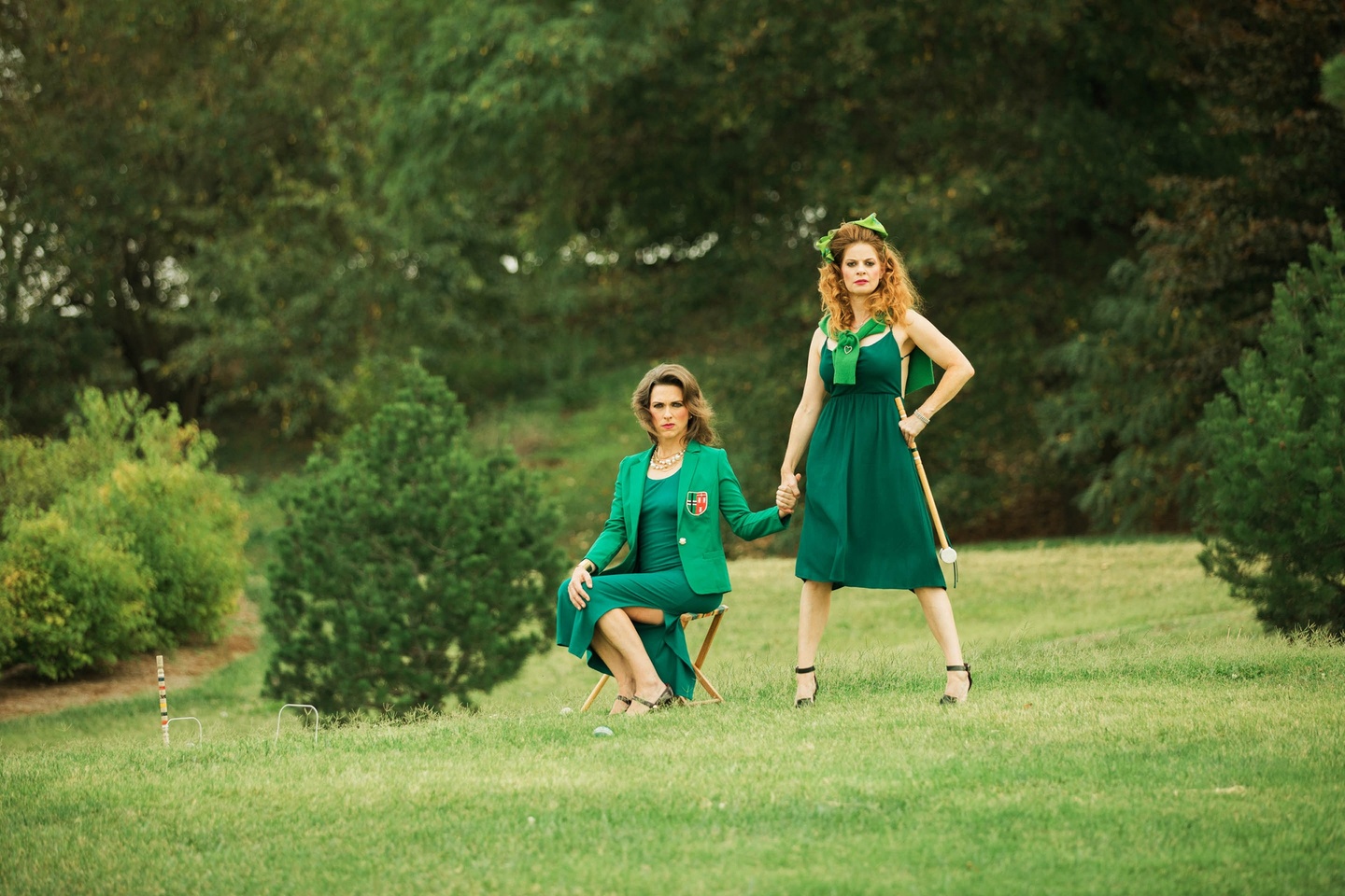A photograph of two people, both in green dresses: the person to the left is seated on a chair, holding hands with the person to the right, who stands in a defiant pose, their left hand on their waist. Their outerwear is tied in a knot and draped on their shoulders. Both people look directly at the camera. In the background, a grassy green field and more bushes and trees beyond.
