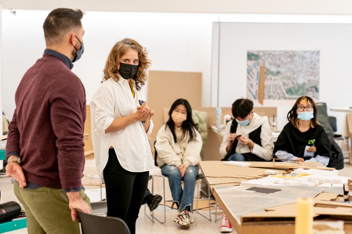 Two instructors stand behind a cardboard city plan model and speak to a room full of students.