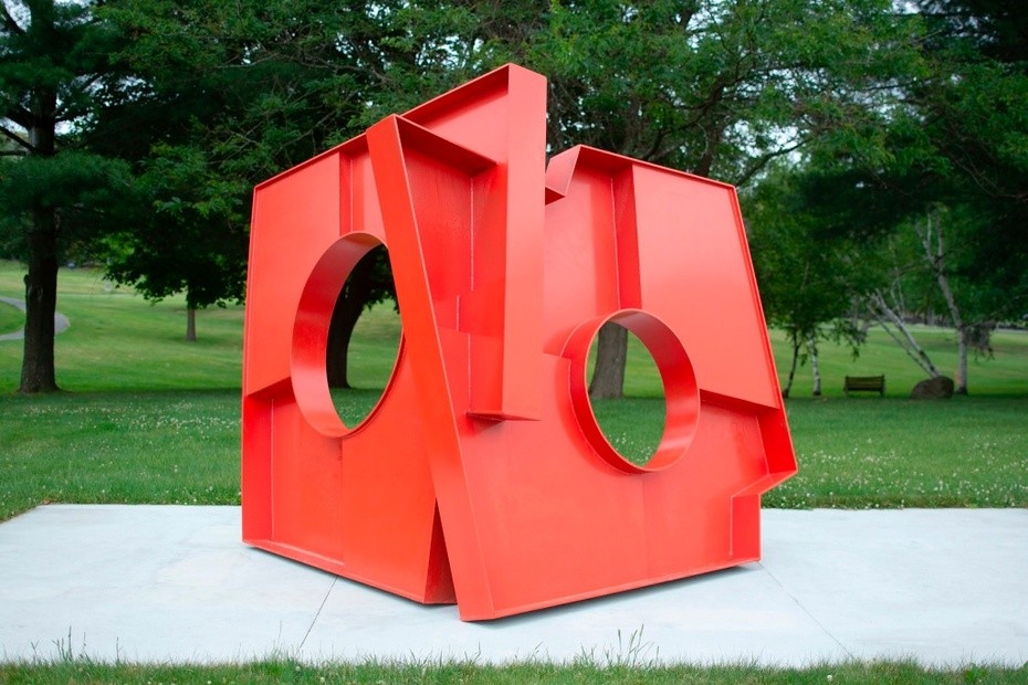 A large red geometric sculpture sitting in the grass on a concrete platform in front of a gray triangular building.