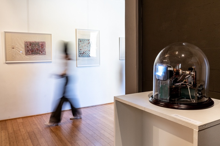 Motion-blurred person moves through a gallery space. In the foreground is a small cathode-ray tube construction housed under a glass dome. 