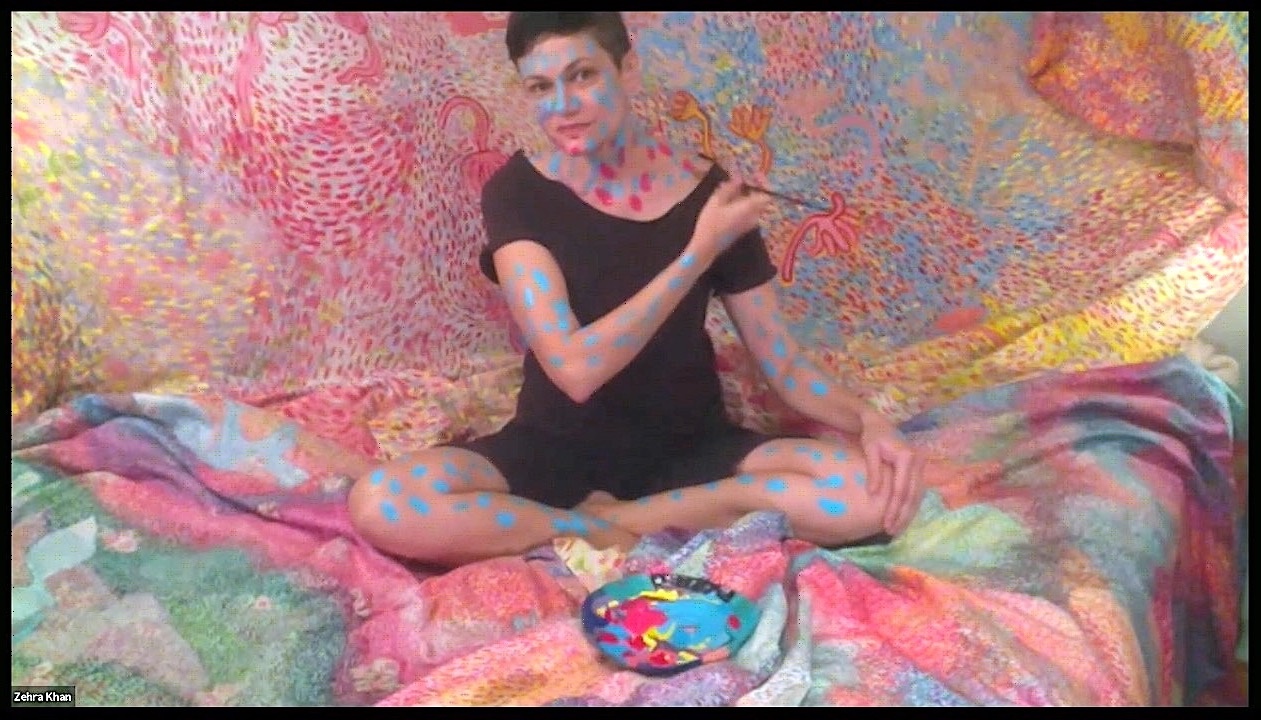 A person wearing a black t-shirt and shorts sits in front of and on colorfully painted fabric while painting their own exposed skin with blue and red dots.