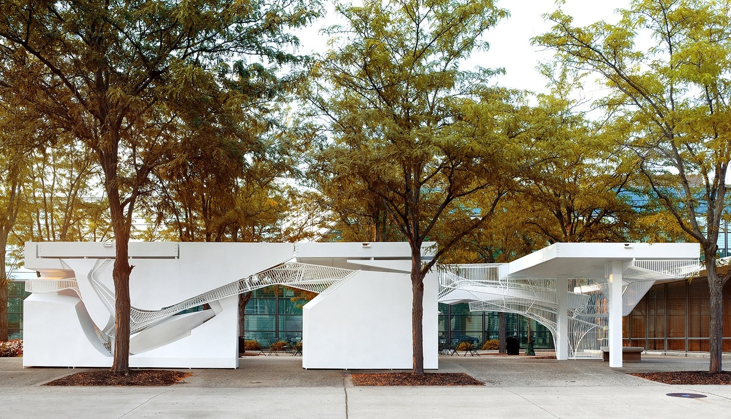 White pavilion installation interwoven through a few trees in an open plaza.