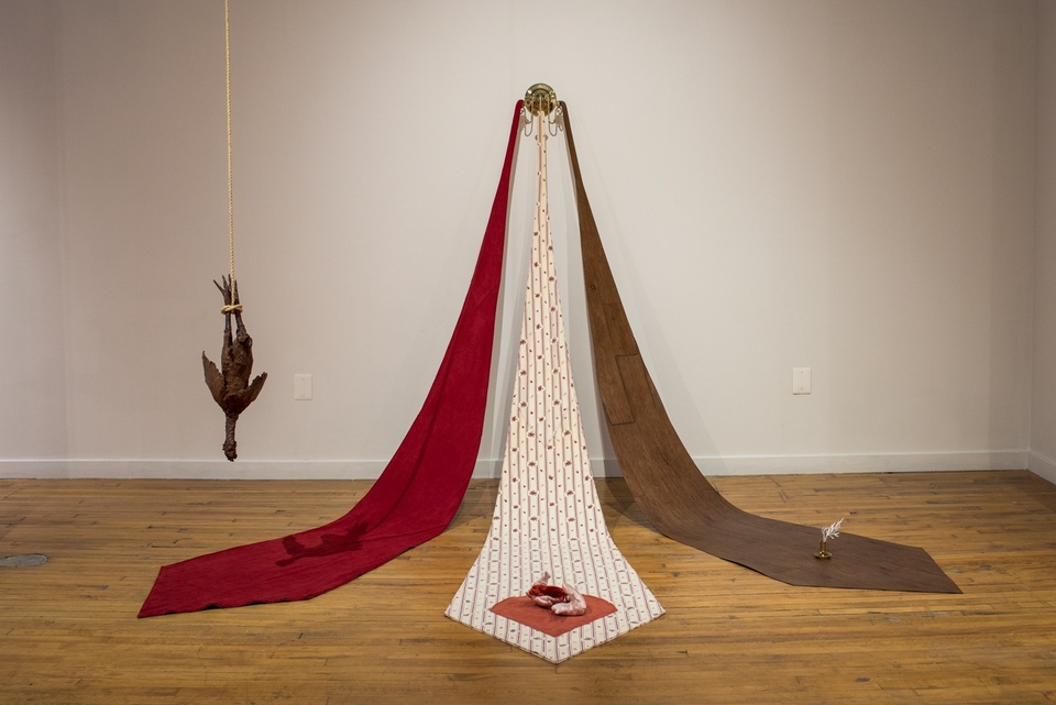 Three oversized necktie-shaped pieces of fabric in red, cream patterened, and brown are draped outward from a sconce on the wall. Each has a mysterious object placed with it.