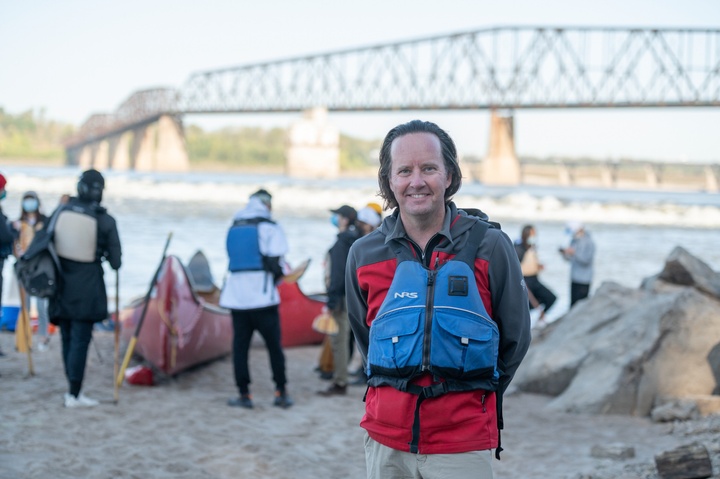 Smiling person in lifevest stands on a riverbank with a bridge behind.