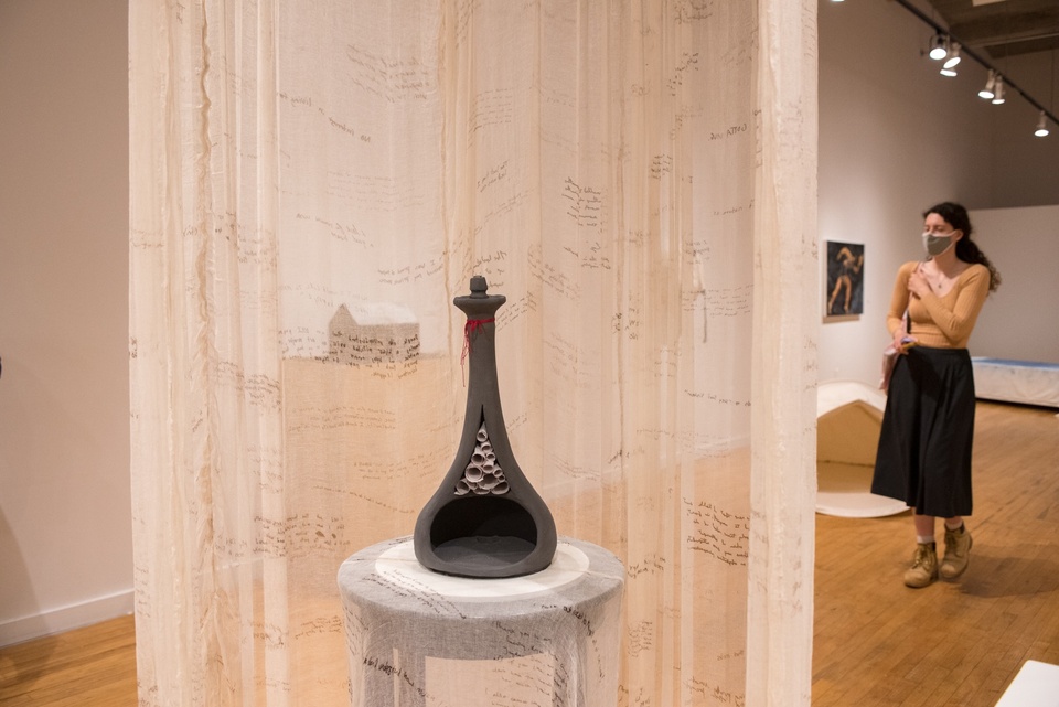 A grey clay vase sliced in half in a vertical cut sits on a stool draped in gauze. A handful of small paper scrolls have been stuffed up into the neck of the vase. A curtain of gauze hangs around the display with spidery writing on it. 