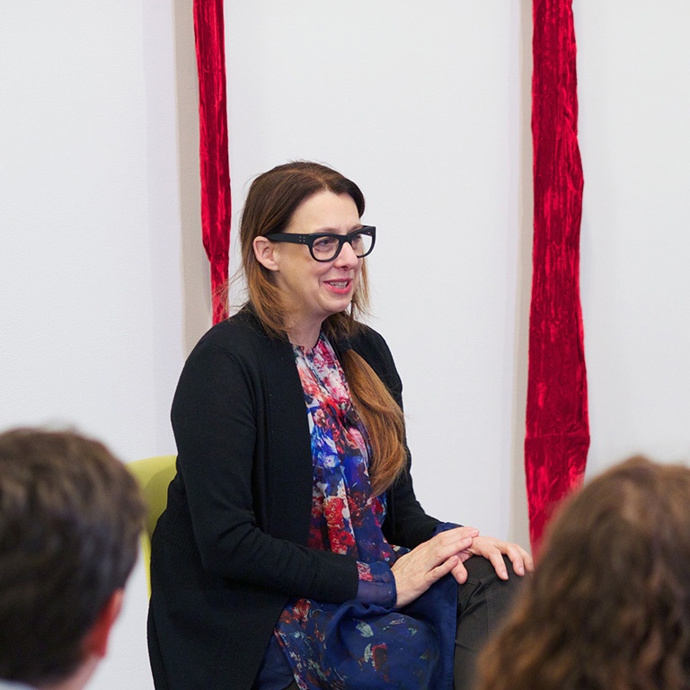Close-up photograph of Beverly Semmes, a white female artist, smiling in front of a crowd with two pieces of long, red velvet visible in the background against the wall.