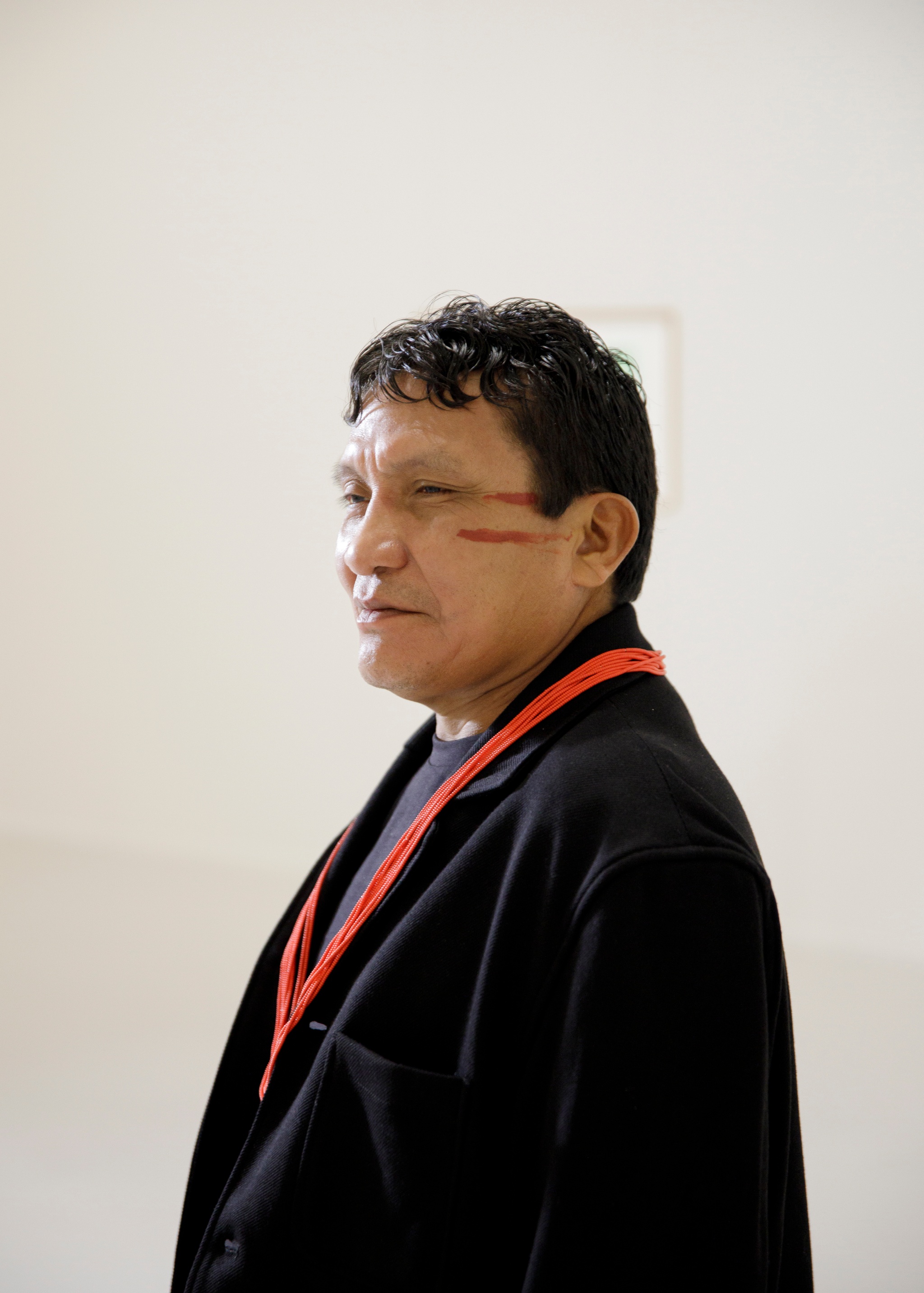 A portrait of artist Joseca Mokahesi. An Indigenous Yanomami man seen in three quarter view in an empty white gallery space. He wears a black blazer with a red ribbon around his neck. He has short dark hair and his face is painted with red lines at the top of his cheekbones.
