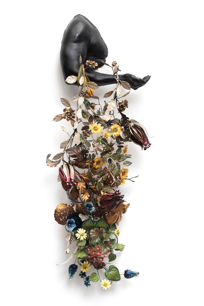 A sculpture by artist Nick Cave with the bust of a figure in profile showing only the shoulder, arm, and part of the chest. The arm holds a garland of flowers that hang down below.