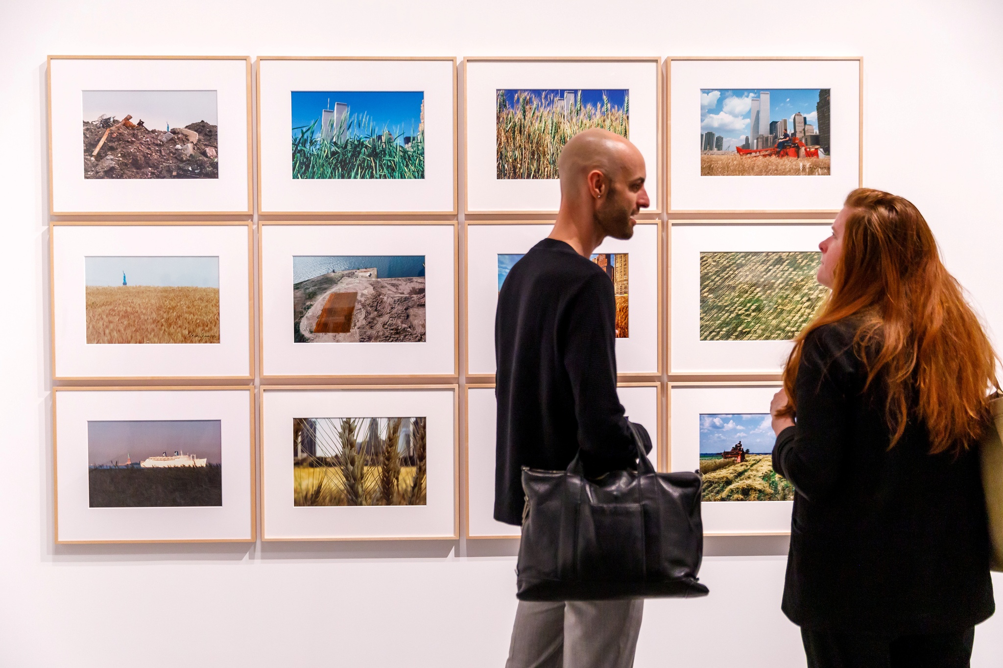 Two people facing each other talking in front of a grid of photographs hung on a gallery wall depicting a wheat field planted by the artist Agnes Denes in Manhattan in 1980