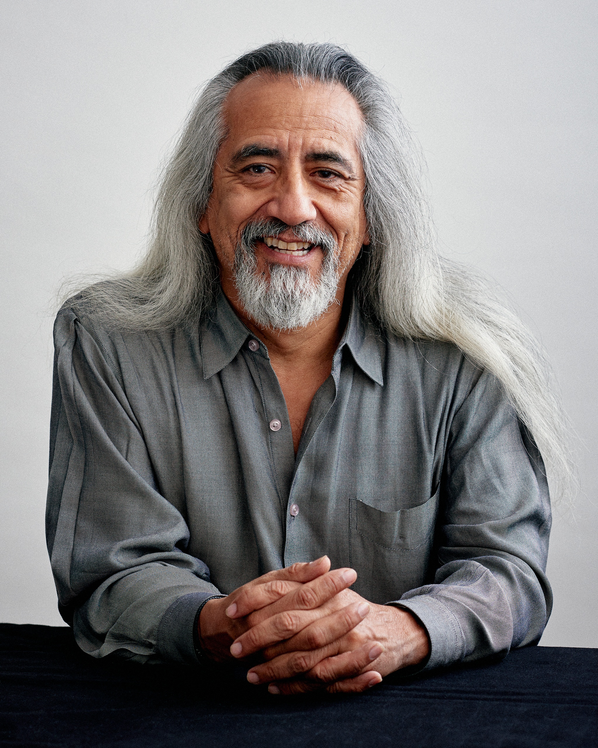 A photo of george emilio sanchez posing with elbows on a table and hands crossed in front of him. Sanchez wears a gray shirt and has long gray hair falling over his shoulders and a gray beard.