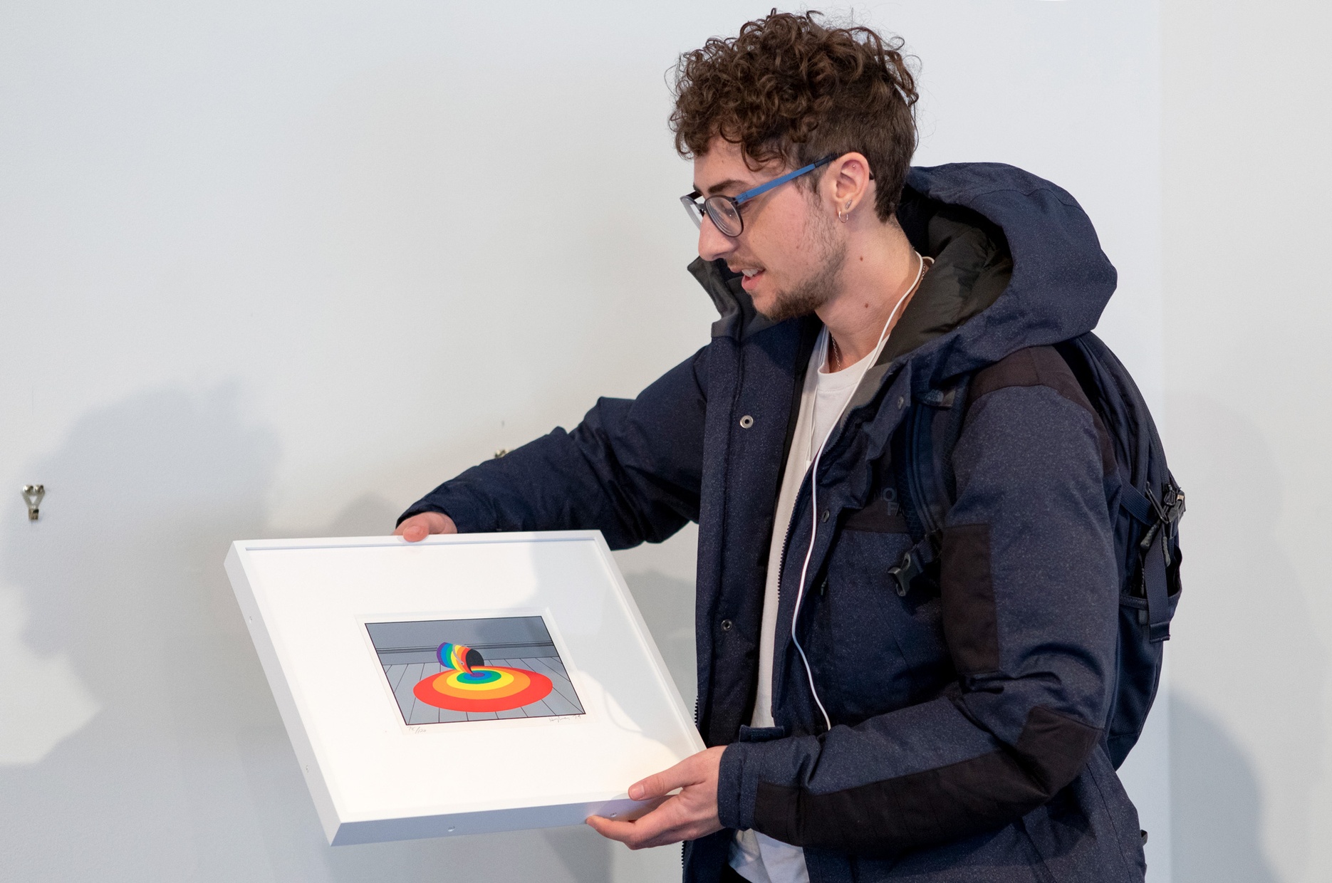 A man is looking down at the framed artwork he is holding by artist Patrick Hughes, which depicts a rainbow paint bucket spilling rainbow paint.